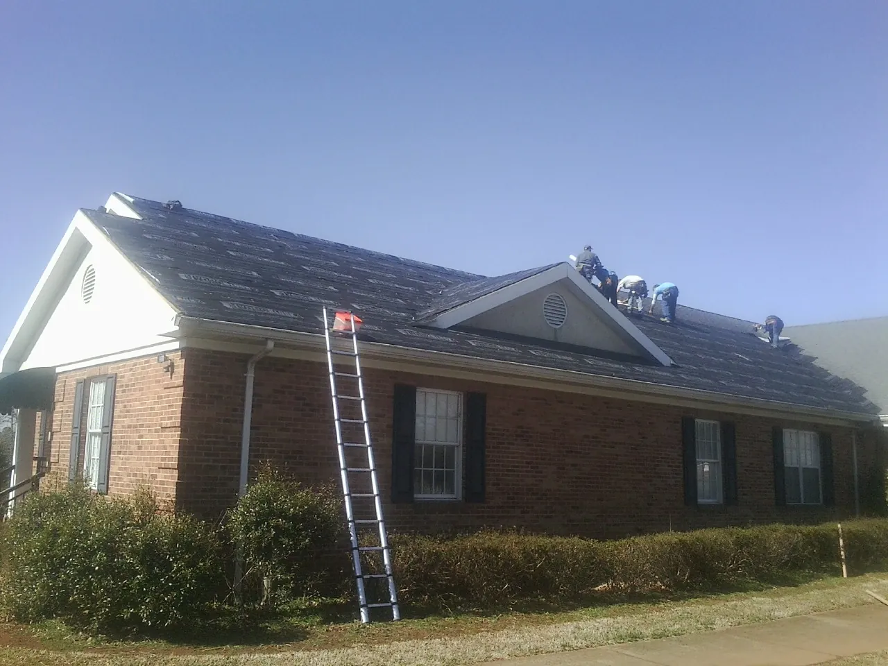 Crew tearing shingles off a brick ranch roof with a ladder set — Emergency Roof Repair work by our crew in Fulshear